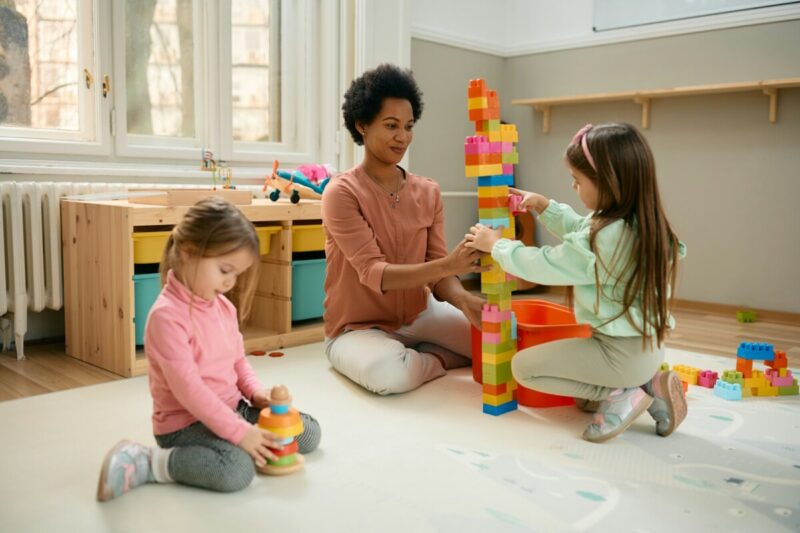 Happy black teacher and preschool kids playing with toy blocks in a playroom.