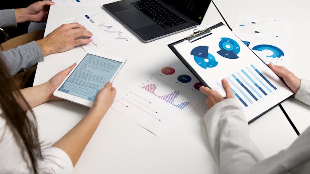 Pessoas utilizando tablet e laptop, analisando gráficos em um ambiente de reunião.